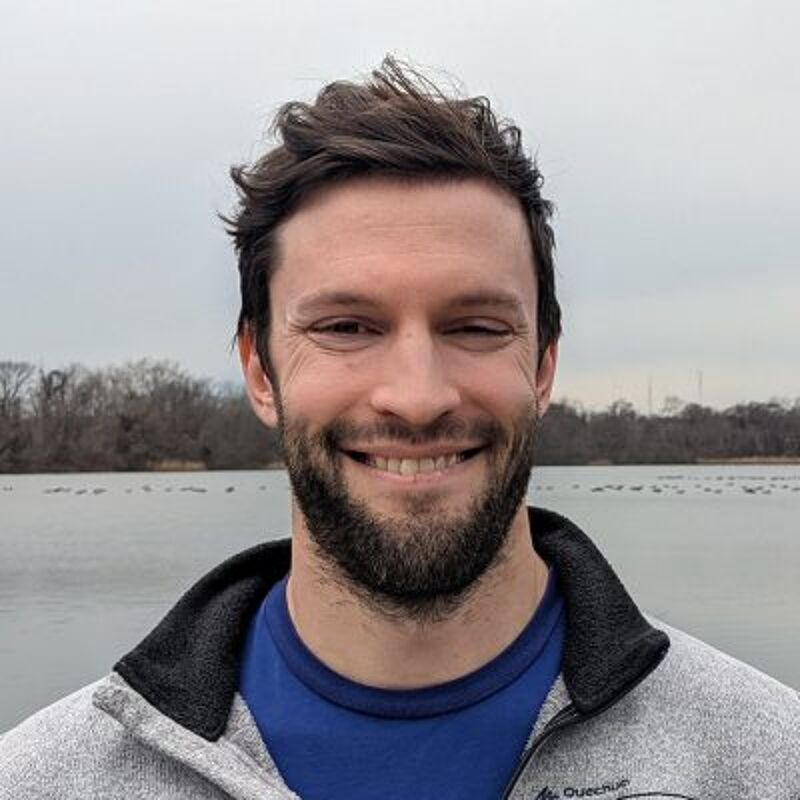 The image shows a man with a beard and short brown hair. He is smiling and looking at the camera. He is wearing a blue shirt and a gray jacket. The background is a lake with some trees and a cloudy sky.